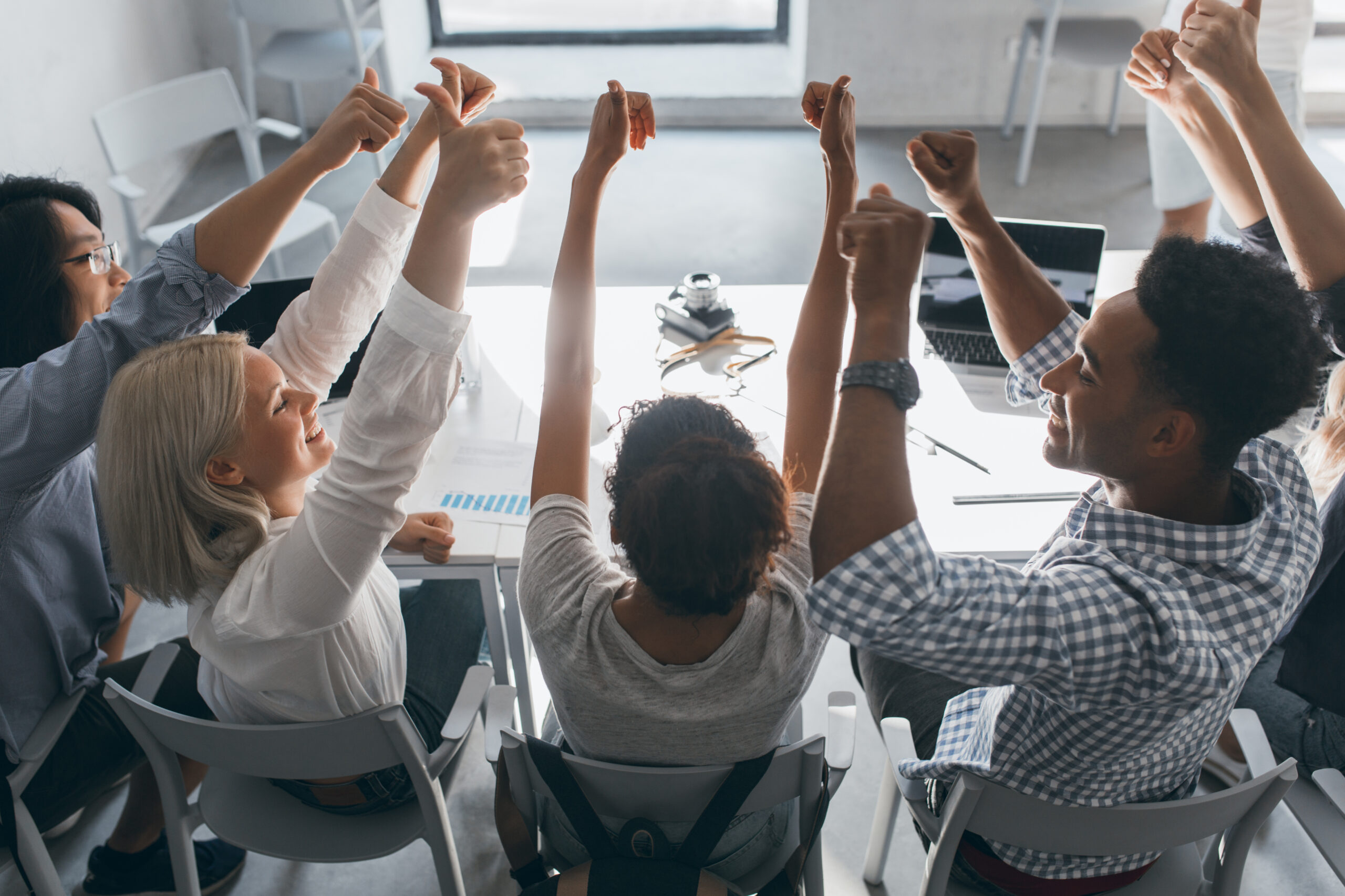 Portrait from back of glad students sitting together at the table and raising hands. Indoor photo of team of freelance specialists having fun after hard work in office.